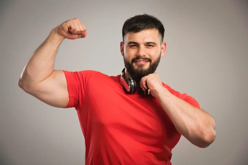 A man in a red shirt and headphones around his neck poses, enhancing his biceps.